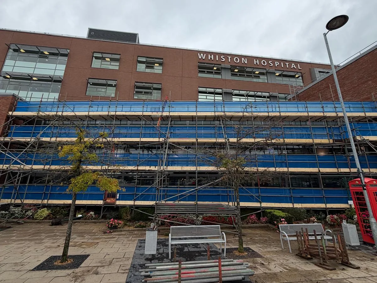 Netted independent scaffold along the glazed atrium of Whiston Hospital, with the hospital entrance behind