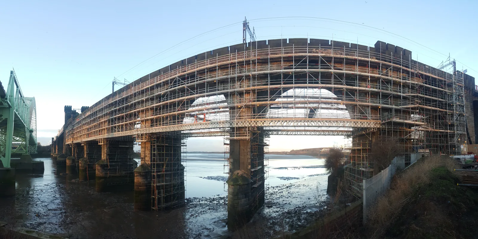 Fully scaffolded rail viaduct spanning a wide river, seen from the bank at low tide